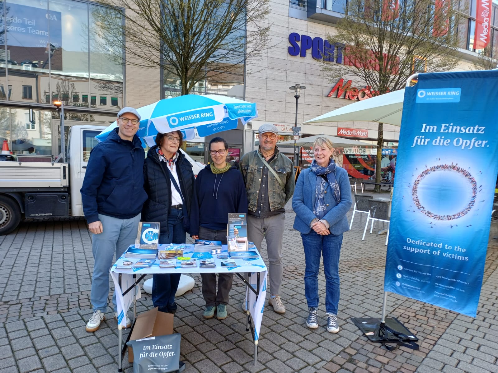 Fünf Mitarbeitende des WEISSEN RINGS stehen vor einem Sonnenschirm und einer Beachflag mit WR-Log, vor Ihnen steht ein Tisch mit Flyern und Infokarten.